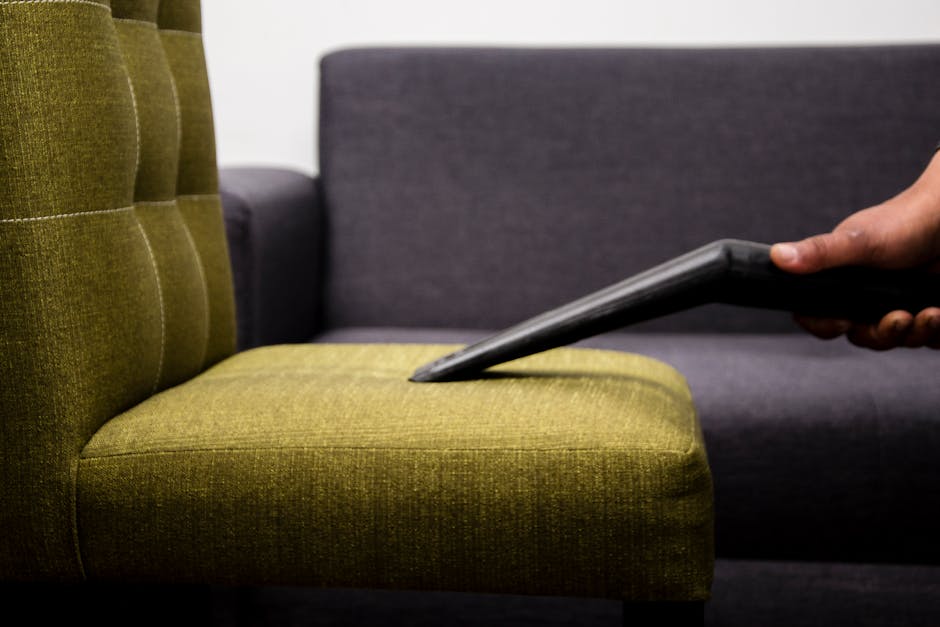 A close-up view of a professional cleaning process on a textured, grey car carpet using a handheld steam or extraction device with a black hose and white nozzle, held by a person wearing a black glove