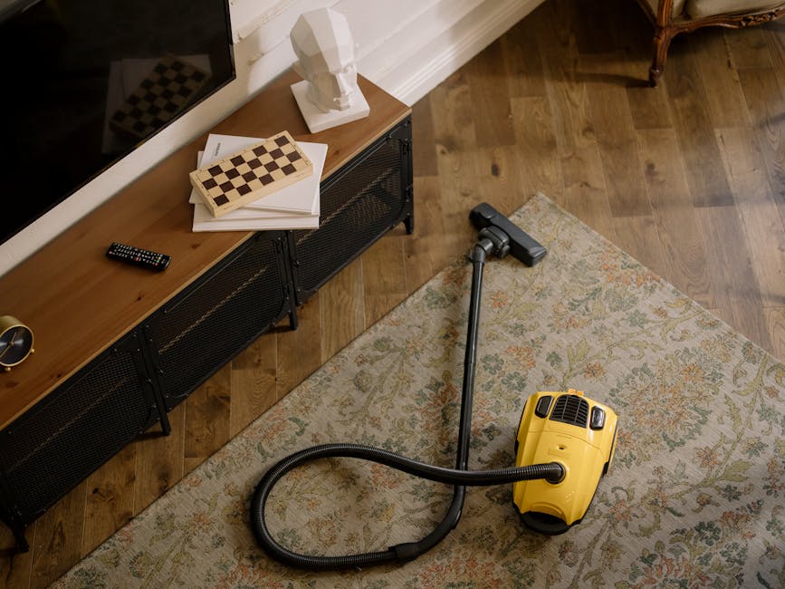 A young boy using a vacuum cleaner to perform surface cleaning and deep cleaning on a beige sofa in a well-lit living room, with an older man resting on the sofa. The living room features hardwood flo